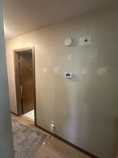 Empty hallway with beige walls, wooden door, wall clock, thermostat, and electrical outlet